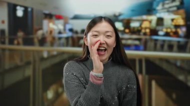 Arka planda canlı mağazalar olan hareketli bir alışveriş merkezinde bağıran, rahat bir kazak giyerken heyecan ve neşe ifade eden bir kadın..