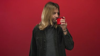 Uzun sarı saçlı, yeşil kadife gömlekli bir adam stüdyoda dudaklarına kırmızı bir kahve fincanı tutuyor..