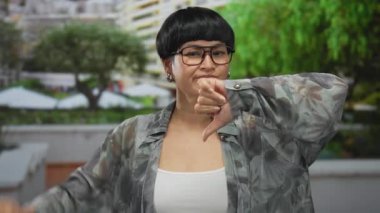 Şehir ortamında gözlük ve çiçek desenli gömlek giyen, yemyeşil ve bulanık arkaplan sahibi, dışardaki kadın baş parmaklarını dışa vuruyor..