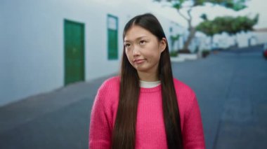 Pembe kazaklı kadın şehir sokaklarında duruyor. Arka planı bulanık. Açık havada düşünceli bir yüz ifadesi sergiliyor..