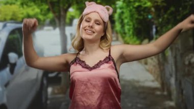 Pembe uyku maskesi takan bir kadın ve açık havada yeşil bir caddede açık havada gülümseyen saten bir kadın. Arka planda bulanık park edilmiş arabalar, rahat bir sabah havası yakalıyor.