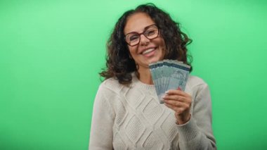 Orta yaşlı bir kadın yeşil bir duvar arkasında Malezya bankaları tutuyor, gülümsüyor ve finansal başarı sergiliyor..