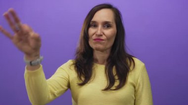Bir kadın gülümsüyor ve mor bir arkaplan karşısında hoş bir jest yapıyor, sarı bir süveter giyiyor, onay ve güven veriyor..