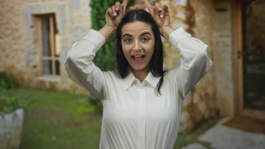 Güneşli bir günde elleri havada, beyaz gömlekli, taşralı bir sokak arkaplanında neşeli bir jest yapan kadın İspanyol gençliğinin neşesini ve doğallığını yakalıyor..