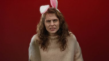 Kahverengi saçlı, tavşan kulağı takan, izole edilmiş kırmızı arka planda rahat süveter içinde kafa karışıklığı gösteren bir kadın.