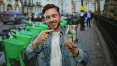 Sakallı ve kot ceketli genç adam şehir caddesinde elinde bir kavanoz dolarla arka planda yeşil scooterlarla baş parmak hareketi yapıyor..