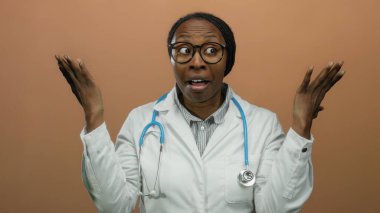 Kadın doktor şaşkınlığını ifade ediyor. Elleri kahverengi arka planda, gözlük ve steteskop takıyor.