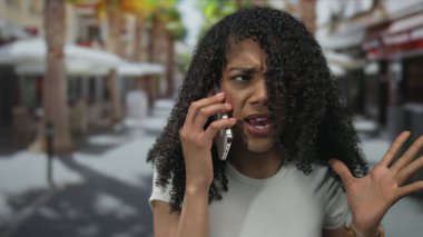 Beyaz tişört giyen Afro-Amerikalı kadın şehir sokaklarında güneş ışığı altında el kol hareketi yaparken akıllı telefonu kulağına tutuyor..