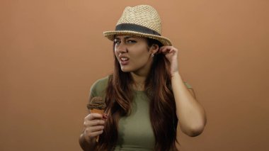 Kafası karışık bir ifade sergileyen kahverengi duvara dondurma tutan şapkalı genç bir kadın.