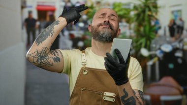 Sakallı, önlüklü ve eldivenli bir adam, şehir sokağındaki dövmeleri görünür olan defteriyle düşünceli bir şekilde el kol hareketi yapıyor ve etrafta sanatsal bir hava yaratıyor.