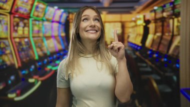 Kapalı alanda gülümseyen bir kadın, canlı bir kumarhane ortamında renkli kumar makinelerini işaret ederek neşeli bir atmosfer yaratıyor..