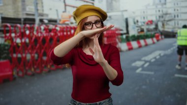 Şantiyede gözlüklü, sarı bereli, parlak kırmızı bariyerli ve bulanık şehir zeminli bir kadın.