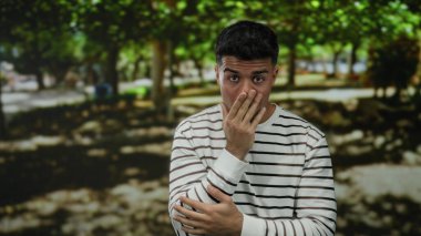 Çizgili tişörtlü İspanyol adam gölgeli ağaçlarla kaplı güneşli bir parkta şaşırmış görünüyor..