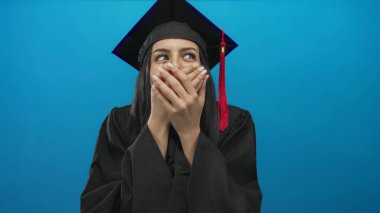 Mezuniyet üniformalı genç İspanyol kadın başarısını kutlayan mavi arka plan duvarı karşısında ağzını şaşkınlıkla kapatıyor..
