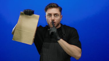 Sakallı ve önlüklü genç bir adam, mavi arka planda bir kesekağıdını sessizce tutuyor. Garson olarak eldiven takıyor, gizli bir jest yaparak paket servisi yapıyor..
