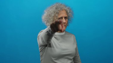 Mavi arka planda ciddi bir ifadeye sahip bir kadın, gri saçlı olgunluğu, orta yaşı ve soyutlanmış ortamları vurguluyor..