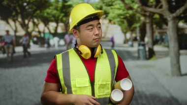 Kentsel caddede güvenlik ekipmanı giymiş genç bir adam elinde planlar, açık bir şehir ortamında mimari planlama ve inşaatı sembolize eden dostane bir jest sunuyor..