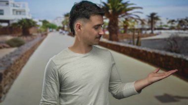 Genç ispanyol adam neşeli bir deniz kenarında yürüyüş yolunda palmiye ağaçlarıyla neşeli bir şekilde el kol hareketi yapar, açık deniz kenarında günlük açık süveter giyer.