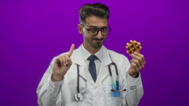 Steteskoplu genç İspanyol doktor mor stüdyoyu işaret eden waffle ve parmak tutuyor; sağlıklı mizah.