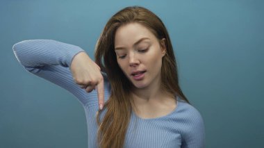 Mavi süveter giyen genç beyaz bir kadın, parlak mavi bir stüdyoda parmağıyla işaret ediyor; neşeli bir eğlence..