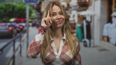 Gündüz vakti işlek bir şehir caddesinde gülümserken, uzun saçlı sarışın kadın parmağıyla başını işaret ediyor..
