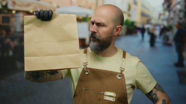 Sakallı bir adam, şehir caddesinde kağıt çanta tutuyor. Eldiven ve önlük takıyor. Gündüz vakti açık havada çok düşünceli görünüyor..