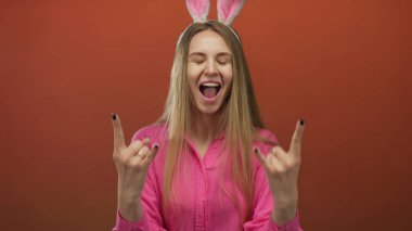 Paskalya tavşanı kulağı takan ve iki eliyle kaya jesti yapan bir kadın, izole edilmiş kırmızı bir arka plana yaslanmış, heyecan ve neşe ifade ediyor..