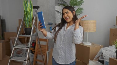 Kadın tablet kullanıyor, evin içinde gülümseyen ve el sallayan Latin kökenli genç bir ev kadını olarak ev hapını arıyor..