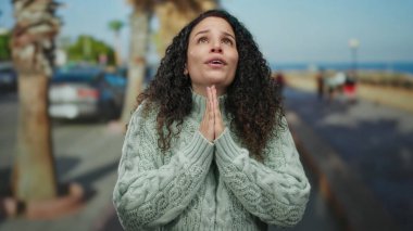 Arabaları ve palmiye ağaçları olan bir sokakta, güneşli bir günde, mavi gökyüzünün altında umut dolu ve huzurlu görünen, rahat bir kazakla dua eden bir kadın..