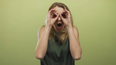 Yeşil bluzlu sarışın kadın, minimalizm tarzında izole edilmiş sarı arka plana karşı şaşırmış bir ifade sergiliyor. Neşeli duyguları, şaşkınlığı ve basitliği vurguluyor..