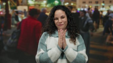 Kalabalık bir sokakta, gözlüklü ve süveterli bir şekilde gülümseyen bir kadın. Etrafı yürüyen insanlarla çevrili, canlı bir şehir atmosferi sağlıyor.