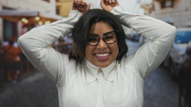 Gözlüklü bir kadın şehir terasında, canlı bir şehir ortamında neşeli bir ifade sergileyen tavşan kulaklı bir jest yapıyor..