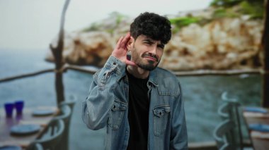 Koyu renk saçlı ve kot ceketli genç adam deniz kenarındaki restoranda poz veriyor bulanık okyanus arkaplanıyla sanki dikkatle dinliyormuş gibi neşeli ifadeler sergiliyor.