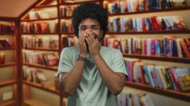 Gözlüklü ve afro saçlı gülümseyen adam, arka planda renkli kitaplarla dolu ahşap raflarla dolu canlı bir kütüphanede duruyor..