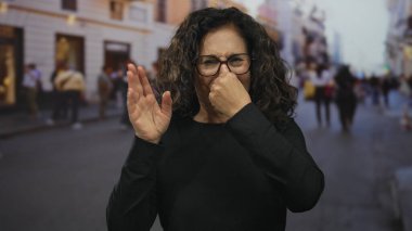 Gözlüklü bir kadın, bulanık binalar ve arka planda insanlarla dolu kalabalık bir caddede tiksindirici bir yüz ifadesi takınarak, etkileyici bir şehir manzarası yaratıyor..