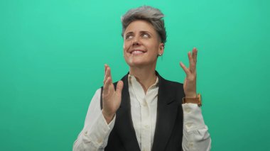 Beyaz gömlekli ve siyah yelekli kısa saçlı kadın, yeşil stüdyoda el çırpıyor ve gülümsüyor; neşe enerjisi kutlamaları ilham kaynağı.