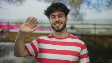 Kırmızı çizgili tişörtlü, sakallı, güneşli bir günde gülümseyen ve el sallayan, bulanık bahçeli genç bir İspanyol.