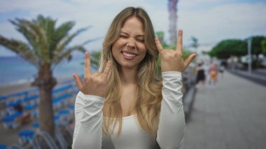 Sarışın kadın, iki eli deniz kenarında kaya boynuzları hareketi yapar; kaya eğlence gençliği enerjisi.