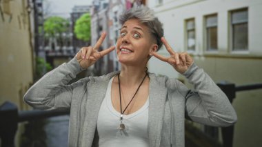 Gri kısa saçlı kadın nehir kanalı kenarında iki eliyle şehir caddesinde barış işareti yapıyor..