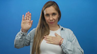 Sol elini göğsüne ve sağ eline koyan bir kadın izole edilmiş mavi bir arkaplana karşı kaldırarak samimiyetini ve bağlılığını sakin ve odaklanmış bir şekilde ifade ediyor..