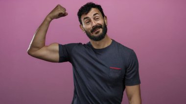 Sakallı İspanyol bir adam canlı pembe arka planda kollarını esnetiyor. Gri bir gömlek giyiyor. Kendine güveni ve gücü var..