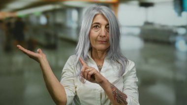 Havaalanı terminalinde gri saçlı yaşlı bir kadın, parmağını göstererek elini beyaz bir gömlekle uzatıyor, eğitici ve bilgilendirici ipuçları veriyor..