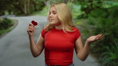 Yeşil parkta kırmızı bir kalp taşıyan, kafası karışık kırmızı tişörtlü bir kadın. Etrafı yemyeşil ağaçlar ve patikalarla çevrili. Sevgi ve kafa karışıklığı taşıyor..