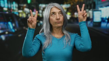 Gri saçlı yaşlı bir kadın, gece aydınlık şehir sokağında jest yapıyor..