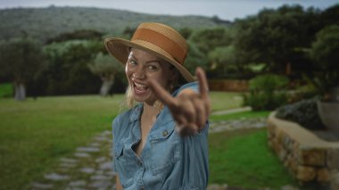 Kot gömlek giyen ve hasır şapka takan kadın yeşil orman açıklığında iki eli kaya boynuzlarını gösteriyor; neşe.