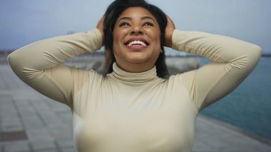 Deniz kenarındaki gezinti güvertesinde güvenle gülümseyen ve saçları şekillendiren bir kadın, açık bir gökyüzünün altında okyanus manzarası gibi neşe ve kendini ifade eden bir sahne..