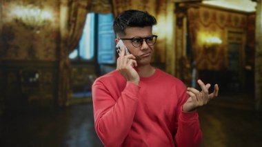 Gözlüklü genç adam lüks otel ortamında telefonla konuşuyor, düşünceli duygularını ifade ediyor..