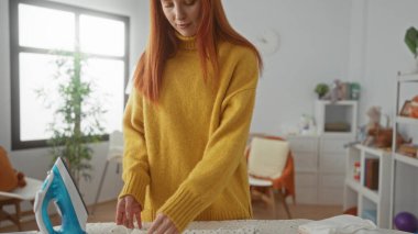 Konforlu ev çamaşırhanesinde kıyafetlerini ütüleyen, sarı kazak giyen, camdan içeri güneş ışığı süzülen ve temiz bir yaşam tarzını ön plana çıkaran bir kadın.