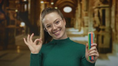 Yeşil kazaklı gülümseyen kadın bir binanın içinde başparmak hareketi yaparken renkli kalemler tutuyor; yaratıcılık.