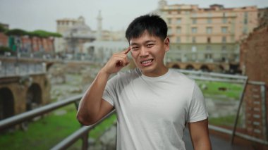 Şaşkın bir hareketi olan genç adam, şehir mimarisi ve yeşilliklerle çevrili hareketli bir caddede duruyor, açık havada şaşkınlığını ifade ediyor..